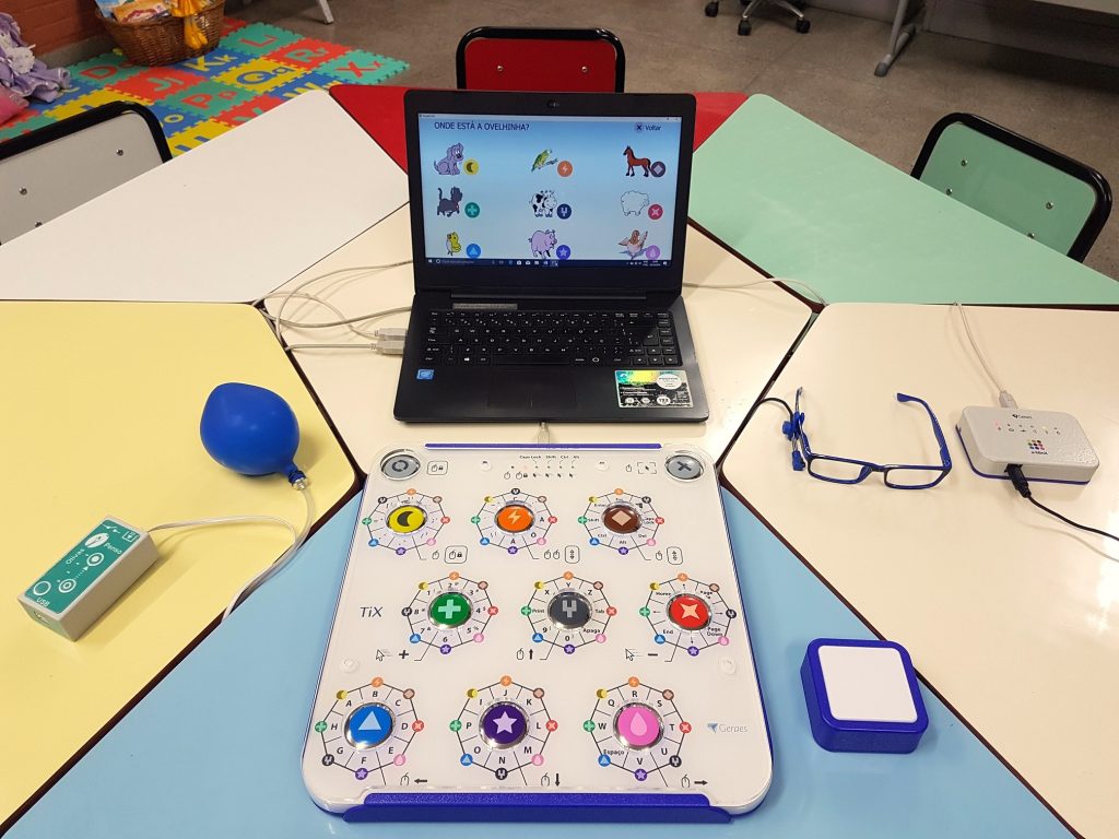 Teclado TiX sobre uma mesa modular de escola, diante de um notebook rodando o software Simplix, cercado pelos acionadores especiais Penso, a-blinX e botão.